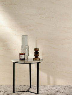 A round table with a marble top holds a white vase, a red candle, and a candle holder with a white candle, set against a wall adorned with Chesapeake Morgana Stone Wallpaper by York Wallcoverings on a gray carpet.