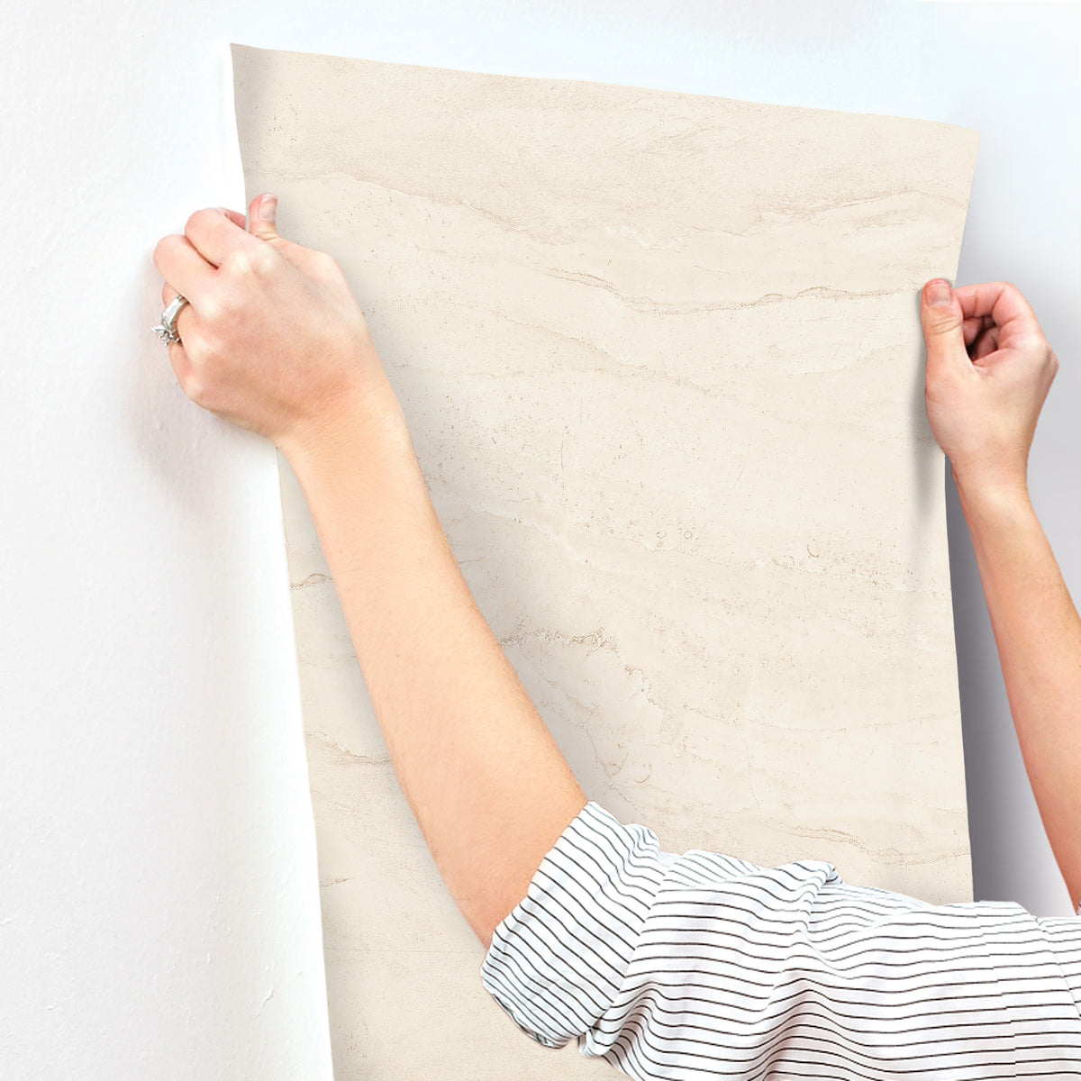 A person in a striped shirt smooths a sheet of Chesapeake Morgana Stone Wallpaper by York Wallcoverings onto a white wall, holding the edges with both hands.