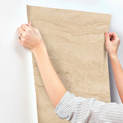 A person in a striped shirt smooths Chesapeake Morgana Stone Wallpaper by York Wallcoverings onto a white wall, holding the top corners with both hands.