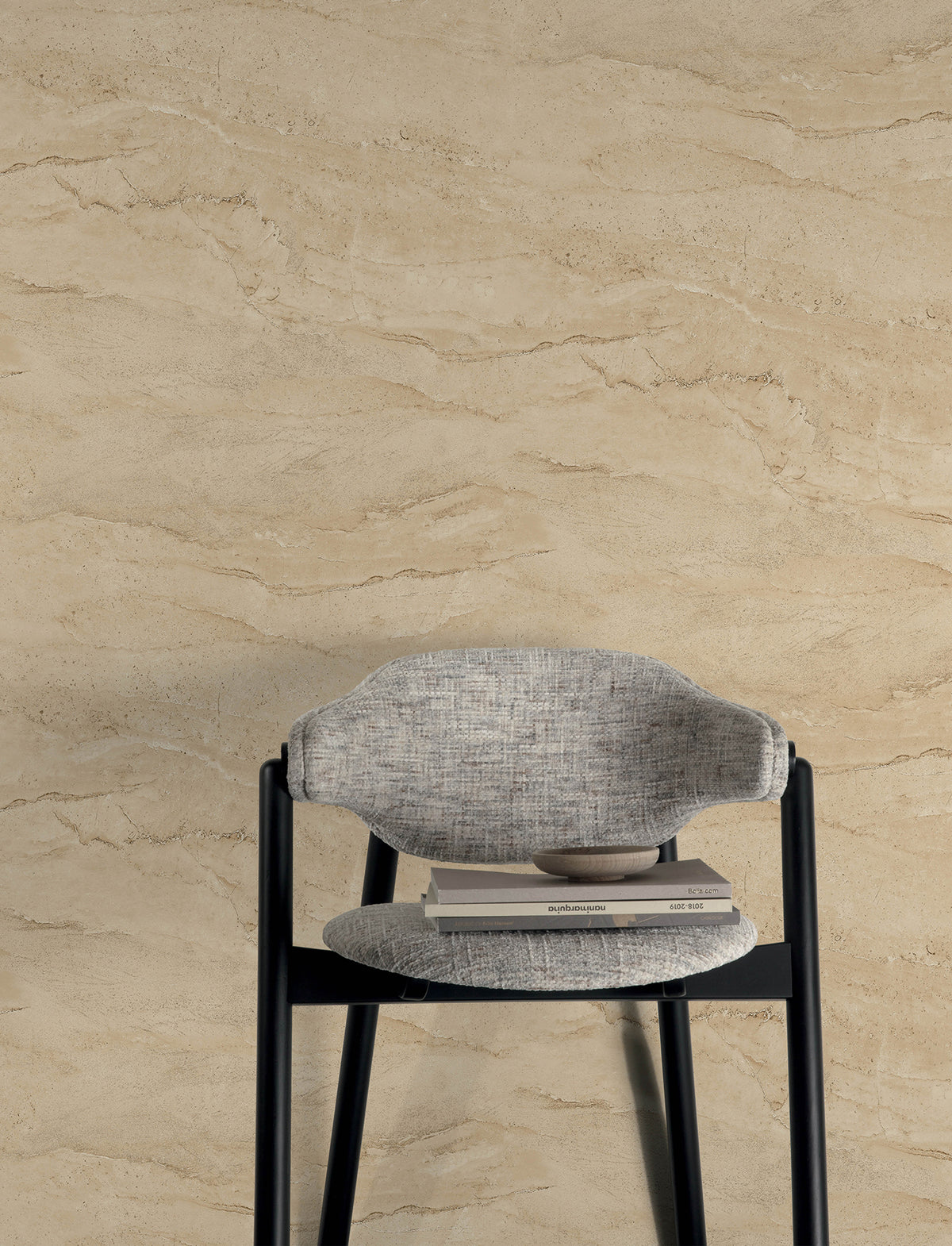 A gray cushioned chair with a bowl and two closed books on the seat is placed against a beige, textured wall covered in York Wallcoverings’ Chesapeake Morgana Stone Wallpaper.