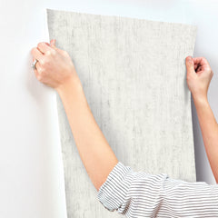 A person wearing a striped long-sleeve shirt is shown holding and aligning a sheet of Chesapeake Ramona Distressed Texture Wallpaper by York Wallcoverings against a white wall; only their arms and hands are visible.