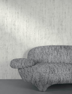A modern, textured gray sofa with rounded shapes sits against a wall covered in Chesapeake Ramona Distressed Texture Wallpaper by York Wallcoverings, partially visible from the left side of the image.
