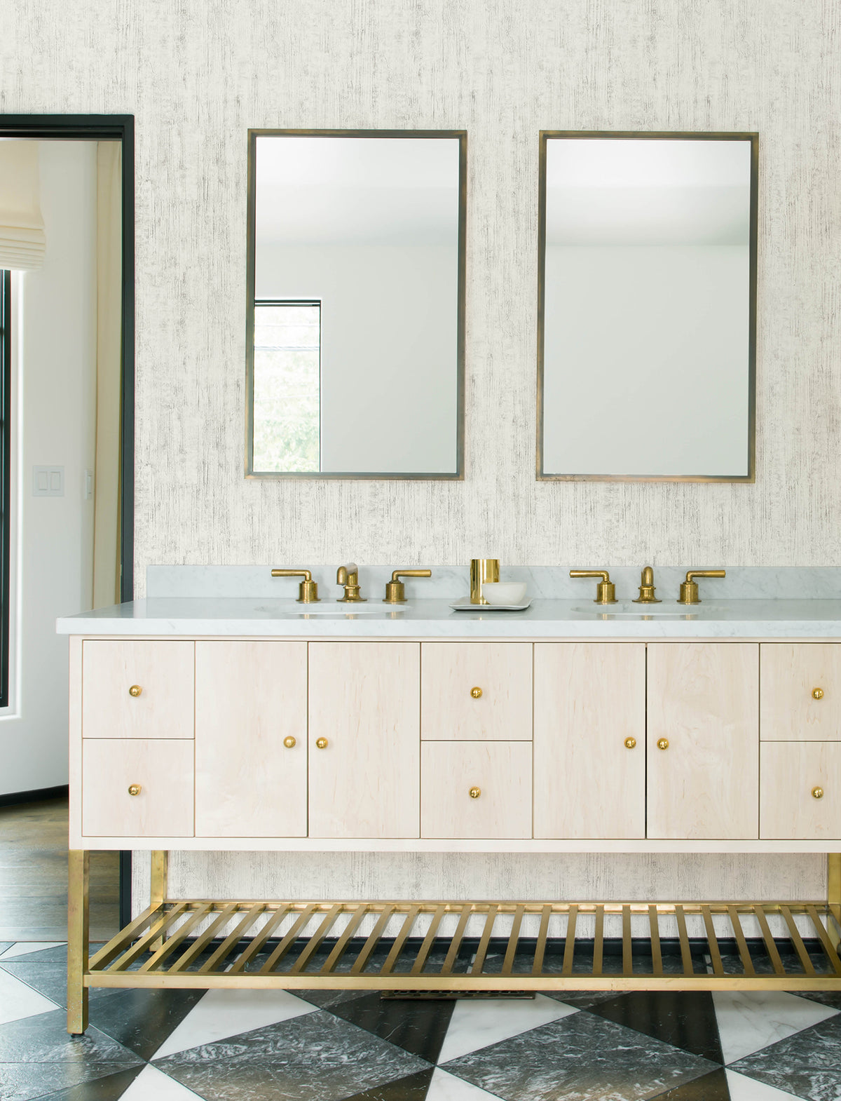 Modern bathroom with two mirrors, double sinks, gold faucets, a light wood vanity, and gold hardware. The floor features a black and white diamond pattern, while the wall is adorned with Chesapeake Ramona Distressed Texture Wallpaper by York Wallcoverings.