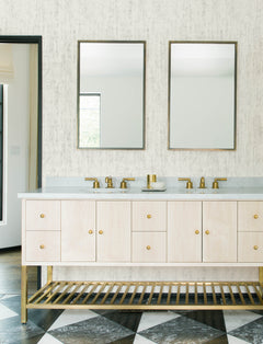Modern bathroom with two mirrors, double sinks, gold faucets, a light wood vanity, and gold hardware. The floor features a black and white diamond pattern, while the wall is adorned with Chesapeake Ramona Distressed Texture Wallpaper by York Wallcoverings.