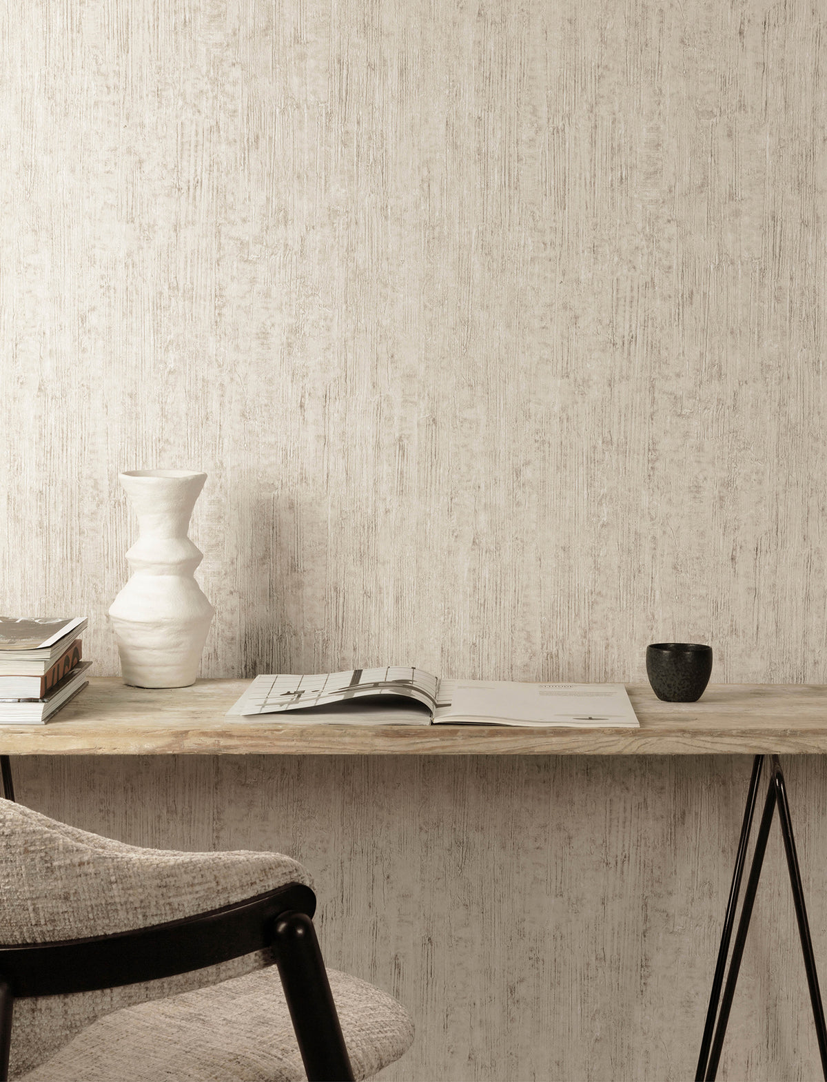 A minimalist desk with a white vase, stacked books, an open magazine, and a small dark cup sits against a wall adorned with Chesapeake Ramona Distressed Texture Wallpaper by York Wallcoverings. A light gray upholstered chair is partially visible in the foreground.