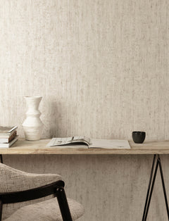 A minimal desk setup features a wall adorned with Chesapeake Ramona Distressed Texture Wallpaper by York Wallcoverings, a ceramic vase, stacked books, an open magazine, and a black cup. A light gray cushioned chair sits at the wooden desk.