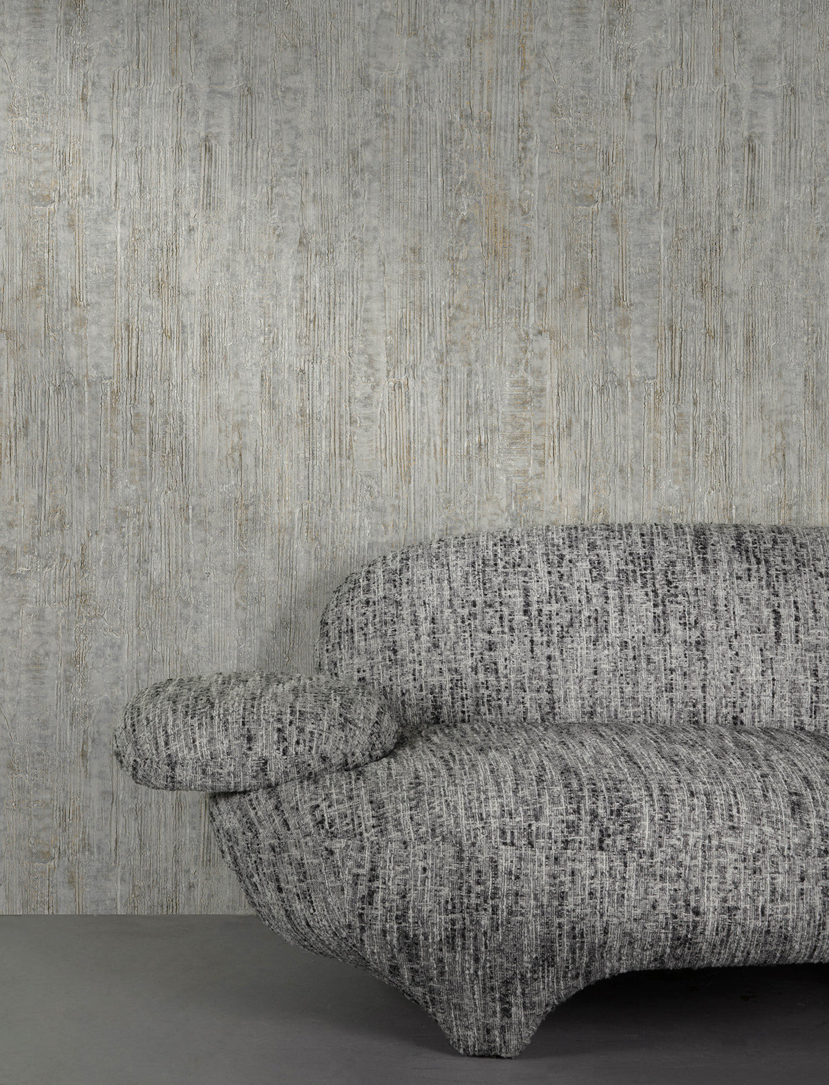 A textured, modern gray sofa is positioned against a wall adorned with Chesapeake Ramona Distressed Texture Wallpaper by York Wallcoverings. Only part of the sofa is visible, showing one armrest and part of the seat and backrest.