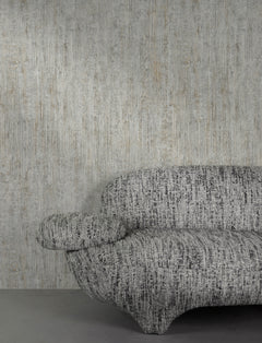 A textured, modern gray sofa is positioned against a wall adorned with Chesapeake Ramona Distressed Texture Wallpaper by York Wallcoverings. Only part of the sofa is visible, showing one armrest and part of the seat and backrest.