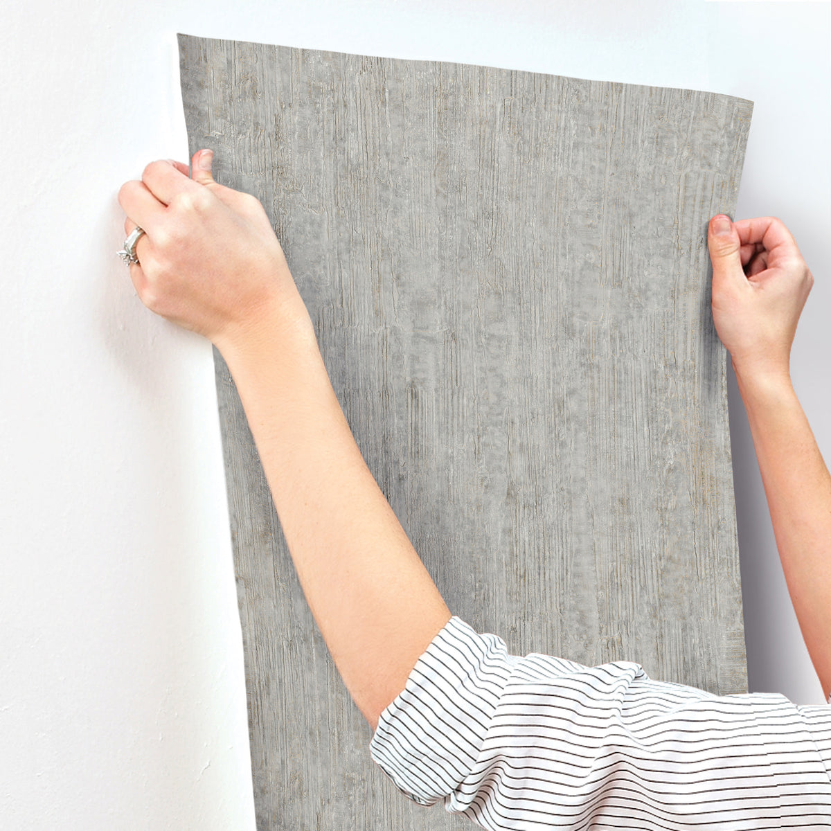 A person applies Chesapeake Ramona Distressed Texture Wallpaper by York Wallcoverings to a white wall, using both hands to hold the gray textured wallpaper in place.