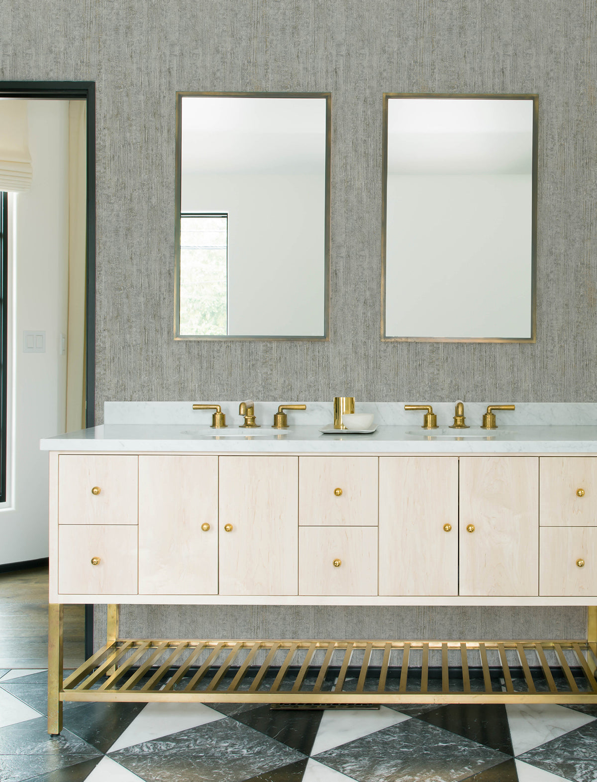 Modern bathroom with two mirrors, gold faucets, and a light wood vanity with gold accents. The floor features a black and white geometric tile pattern, while the walls are adorned with Chesapeake Ramona Distressed Texture Wallpaper by York Wallcoverings in a neutral tone.