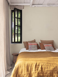 Cozy bedroom with a mustard yellow bedspread, brown and patterned pillows, Chesapeake Elijah Texture Wallpaper by York Wallcoverings on the walls, a rustic wooden bench, open black-framed window, and light curtains letting in natural light.