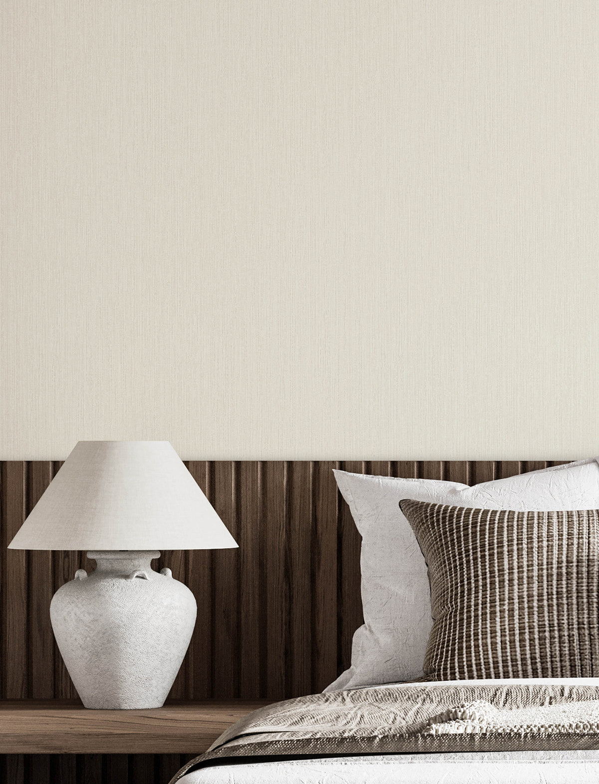 A minimalist bedroom featuring a wooden headboard, a white table lamp, and neutral-colored pillows and bedding, with the wall behind adorned in York Wallcoverings’ Chesapeake Elijah Texture Wallpaper.
