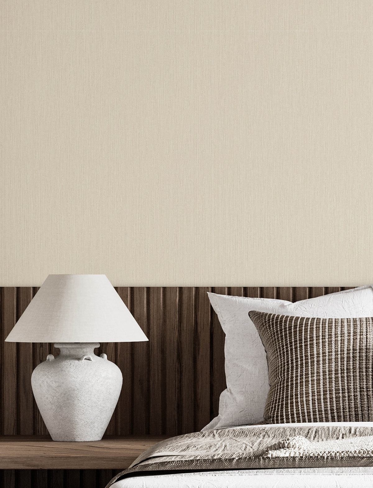 A white table lamp sits on a wooden headboard next to a bed with white and brown striped pillows, with Chesapeake Elijah Texture Wallpaper by York Wallcoverings covering the wall in the background.