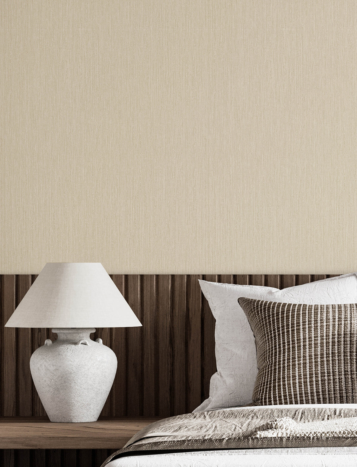 Minimalist bedroom scene featuring a Chesapeake Elijah Texture Wallpaper by York Wallcoverings in beige, complemented by a wooden headboard, a white ceramic lamp, and neatly arranged pillows in neutral tones on a made bed.