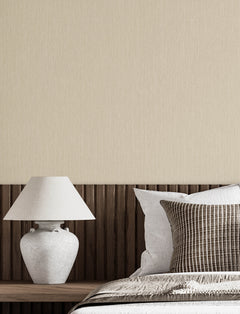 Minimalist bedroom scene featuring a Chesapeake Elijah Texture Wallpaper by York Wallcoverings in beige, complemented by a wooden headboard, a white ceramic lamp, and neatly arranged pillows in neutral tones on a made bed.