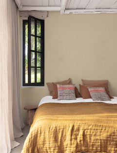 A cozy bedroom featuring a mustard yellow bedspread, brown and patterned pillows, a wooden bench, beige curtains, and walls adorned with Chesapeake Elijah Texture Wallpaper by York Wallcoverings, with natural light streaming through the window.