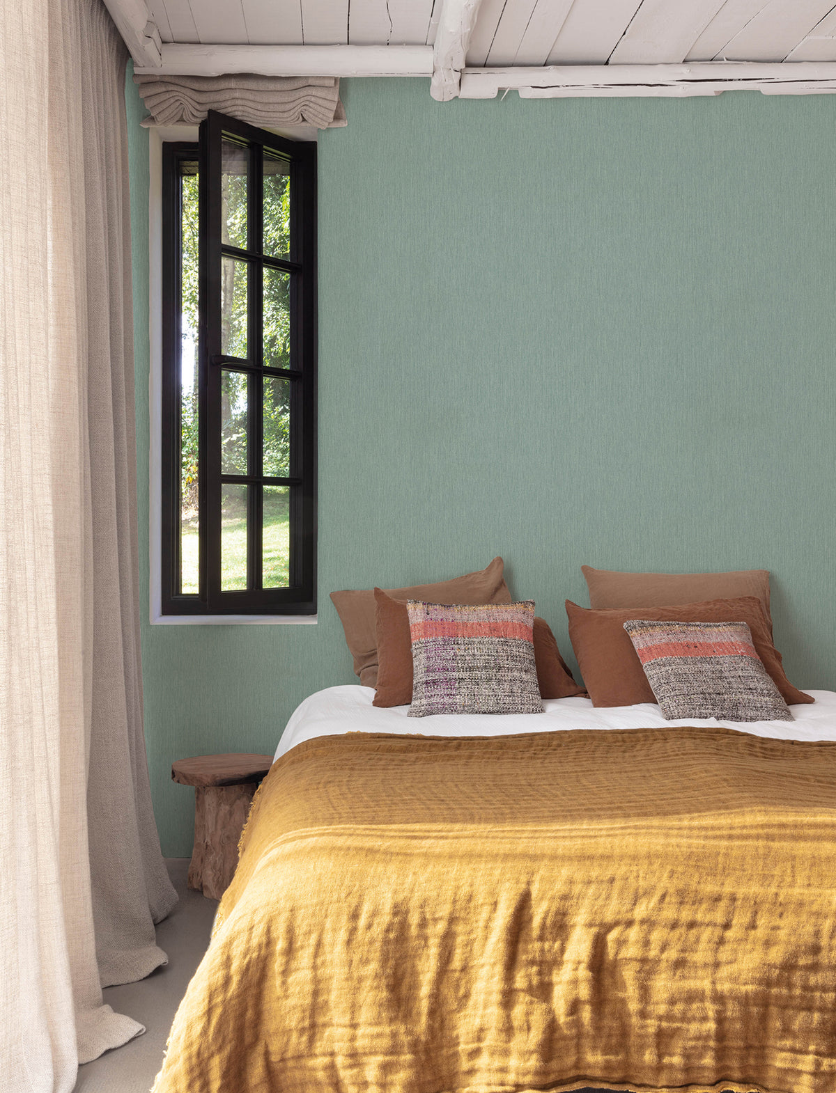 A cozy bedroom featuring an accent wall adorned with York Wallcoverings’ Chesapeake Elijah Texture Wallpaper in green, a window with black frames and beige curtains, a bed topped with brown and red pillows and a mustard yellow blanket. Sunlight filters through the window, illuminating a small wooden bench beside the bed.