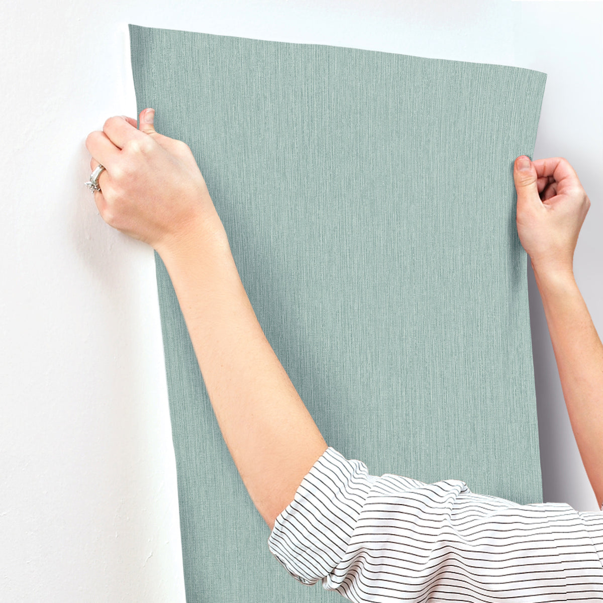 A person wearing a striped shirt is holding up a sheet of Chesapeake Elijah Texture Wallpaper by York Wallcoverings in light blue against a white wall, preparing to install it.