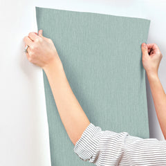 A person wearing a striped shirt is holding up a sheet of Chesapeake Elijah Texture Wallpaper by York Wallcoverings in light blue against a white wall, preparing to install it.