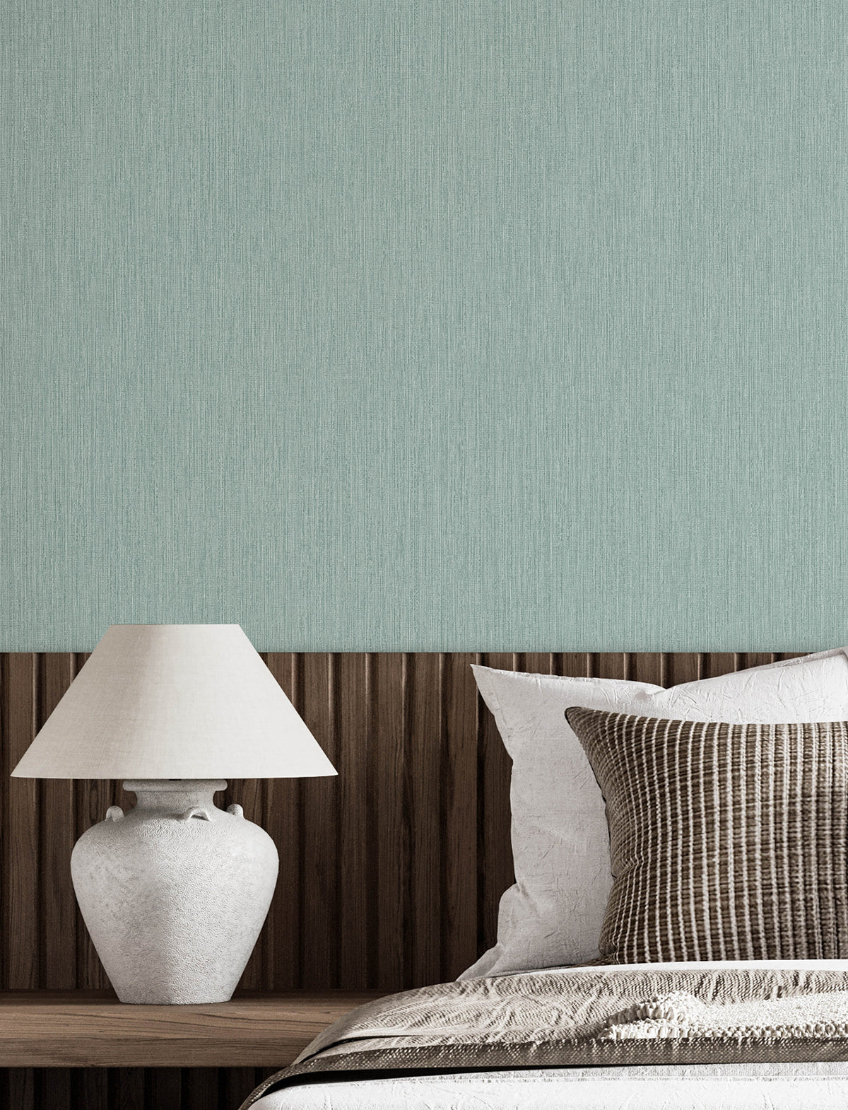 A neatly made bed with white and striped pillows sits next to a wooden headboard and a white ceramic lamp, set against a light green wall adorned with Chesapeake Elijah Texture Wallpaper by York Wallcoverings.