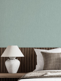 A neatly made bed with white and striped pillows sits next to a wooden headboard and a white ceramic lamp, set against a light green wall adorned with Chesapeake Elijah Texture Wallpaper by York Wallcoverings.
