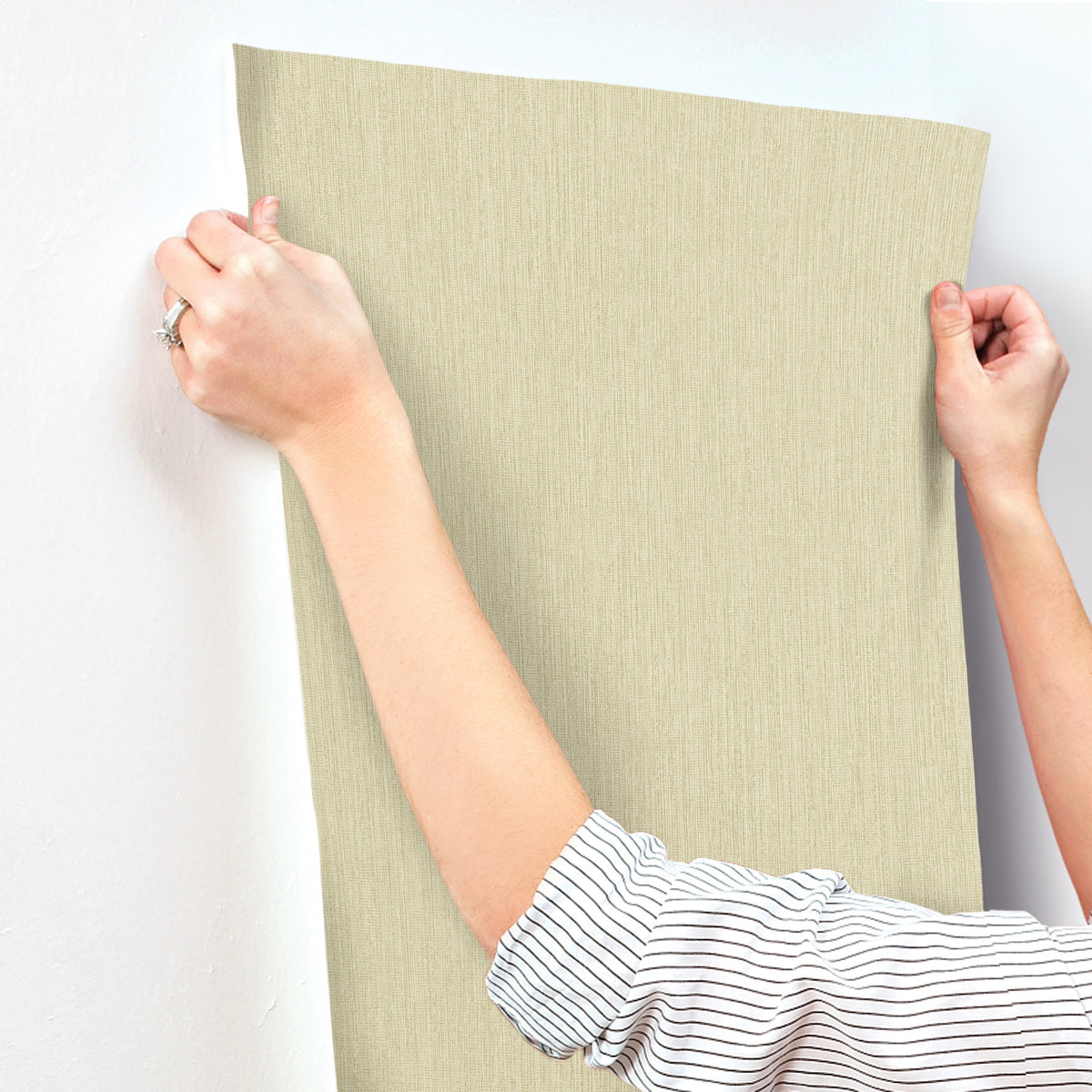 A person wearing a striped shirt hangs Chesapeake Elijah Texture Wallpaper by York Wallcoverings on a white wall, smoothing it with both hands.