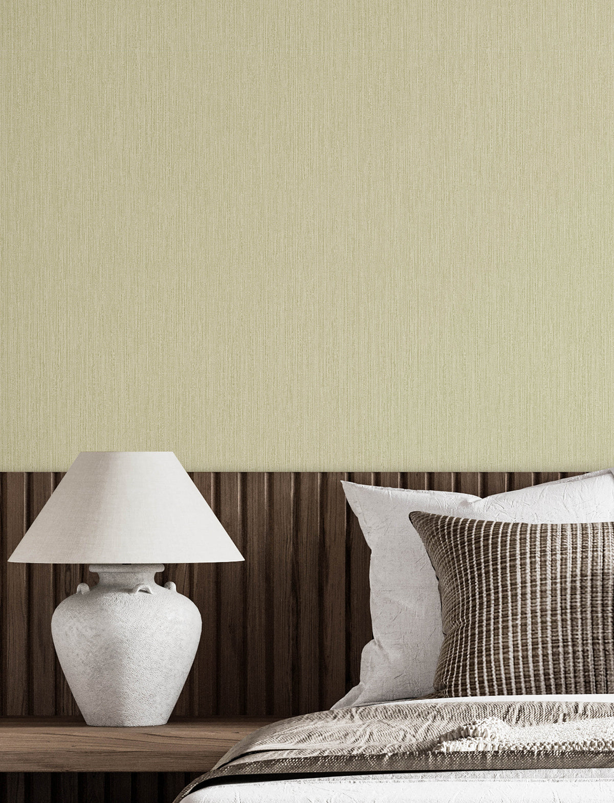 A modern bedroom featuring a white lamp on a wooden nightstand, a bed adorned with white and patterned pillows, and a textured beige wall in the background covered with Chesapeake Elijah Texture Wallpaper by York Wallcoverings.