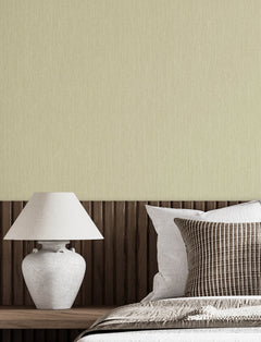 A modern bedroom featuring a white lamp on a wooden nightstand, a bed adorned with white and patterned pillows, and a textured beige wall in the background covered with Chesapeake Elijah Texture Wallpaper by York Wallcoverings.