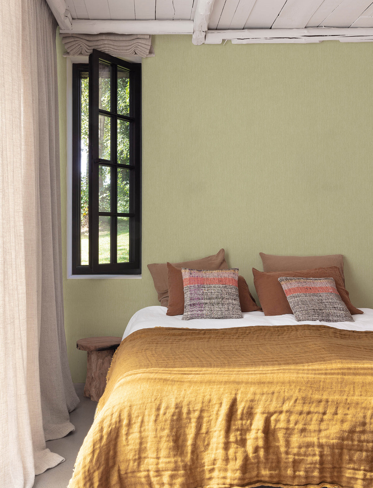 A cozy bedroom featuring Chesapeake Elijah Texture Wallpaper by York Wallcoverings on the walls, a mustard-yellow bedspread, earth-toned pillows, a rustic wood bench, beige curtains, and a black-framed window letting in natural light. The ceiling is accented with white wooden beams.