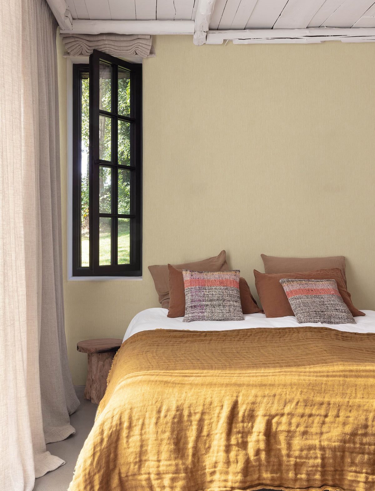 A cozy bedroom featuring Chesapeake Elijah Texture Wallpaper by York Wallcoverings, a mustard yellow bedspread, brown and patterned pillows, a rustic bench, sheer beige curtains, and a black-framed window that lets in natural light.