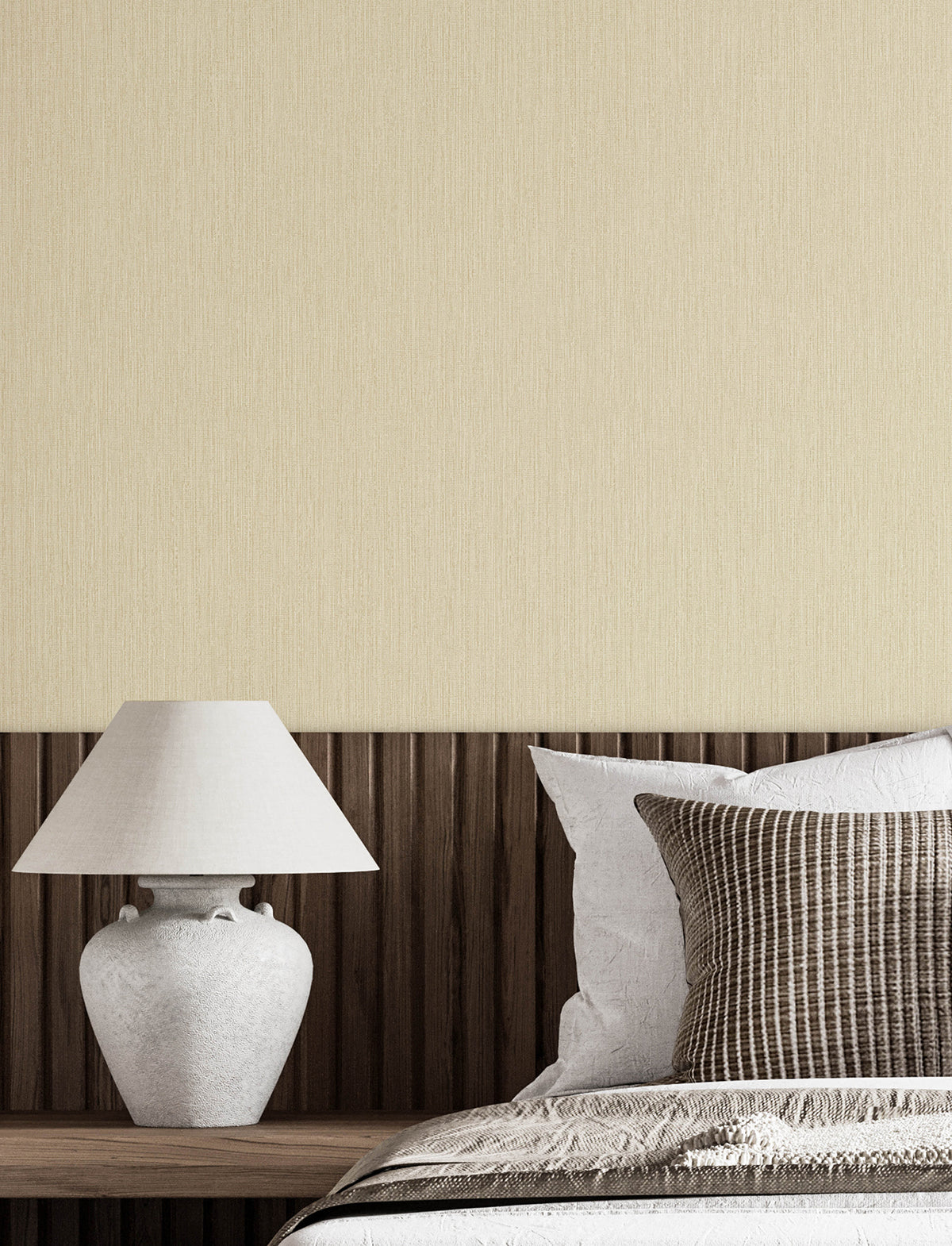 A minimalist bedroom scene featuring a white ceramic table lamp on a wooden nightstand, set beside a bed with neutral-colored pillows and Chesapeake Elijah Texture Wallpaper by York Wallcoverings on the wall behind.