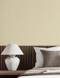 A minimalist bedroom scene featuring a white ceramic table lamp on a wooden nightstand, set beside a bed with neutral-colored pillows and Chesapeake Elijah Texture Wallpaper by York Wallcoverings on the wall behind.