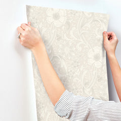 A person in a striped shirt applies York Wallcoverings’ Chesapeake Liora Floral Trail Wallpaper to a white wall, holding the wallpaper at the top corners with both hands.