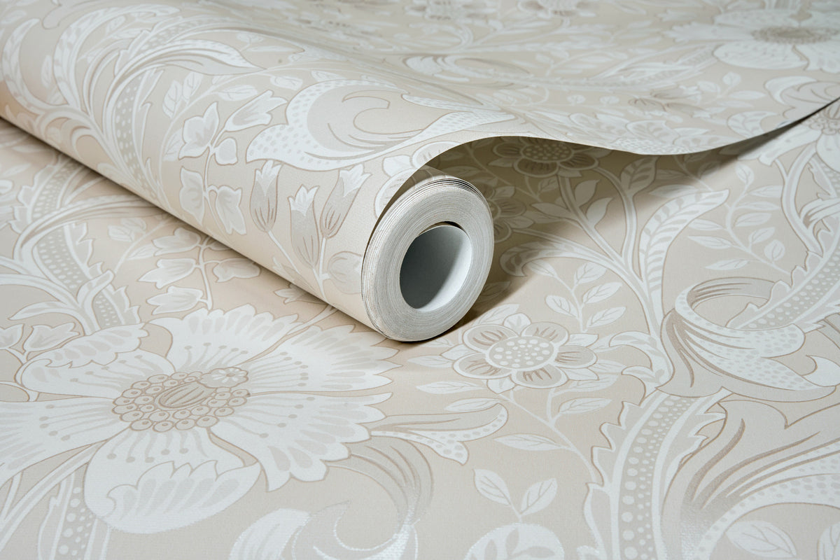 A roll of Chesapeake Liora Floral Trail Wallpaper by York Wallcoverings in light beige, featuring a large, intricate floral pattern, is partially unrolled to display its decorative design on a flat surface.