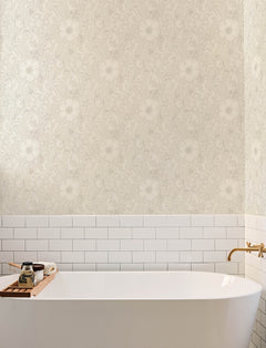 A white bathtub with a wooden tray, gold faucet, white subway tile, and York Wallcoverings’ Chesapeake Liora Floral Trail Wallpaper in beige above.