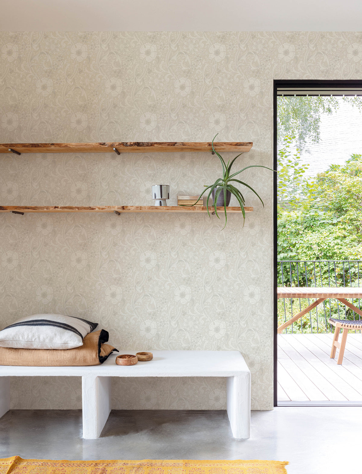 A minimalist room featuring York Wallcoverings’ Chesapeake Liora Floral Trail Wallpaper, with two wooden shelves displaying a plant and decor, a white bench topped with folded blankets and bowls, and an open door revealing a green patio.