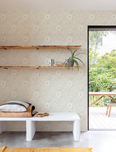 A minimalist room featuring Chesapeake Liora Floral Trail Wallpaper by York Wallcoverings, with a white bench, stacked cushions, wooden shelves displaying a plant and cups, and an open door leading to a patio with greenery outside.