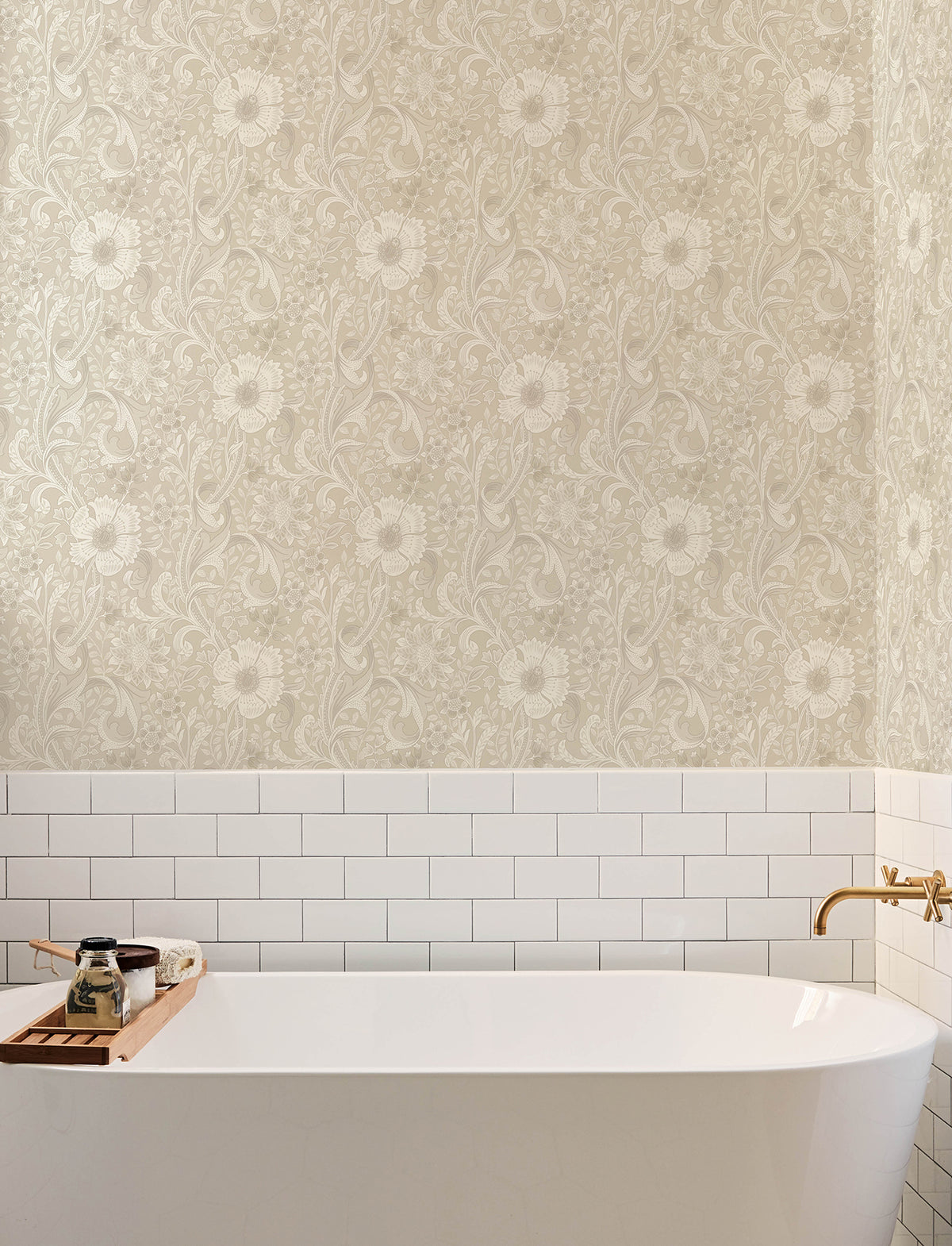 A white bathtub with a wooden tray holding soap and a candle is set against a wall adorned with Chesapeake Liora Floral Trail Wallpaper by York Wallcoverings and white subway tile. A gold faucet is mounted on the wall to the right.