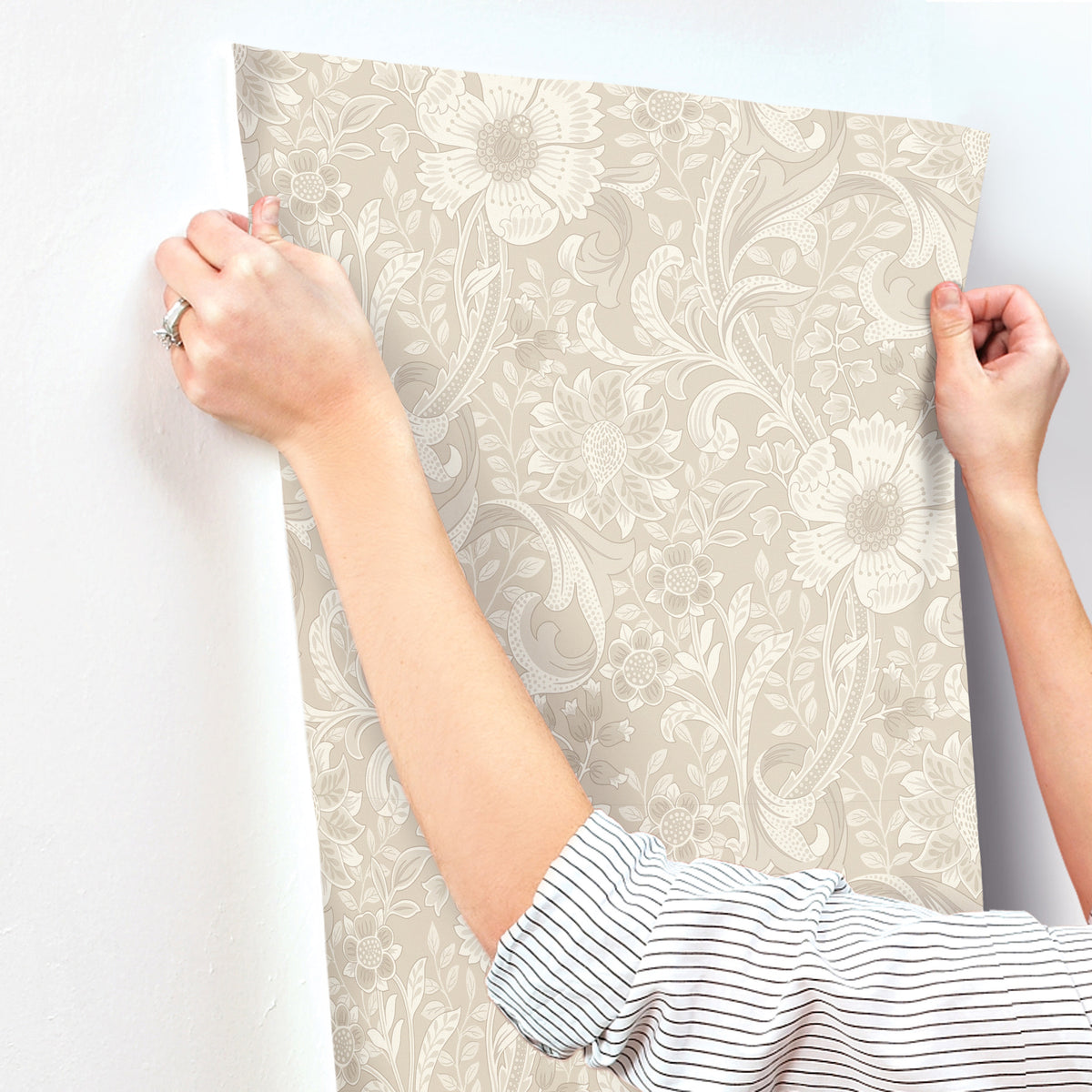 A person in a striped shirt is applying York Wallcoverings Chesapeake Liora Floral Trail Wallpaper to a white wall, smoothing it with both hands.