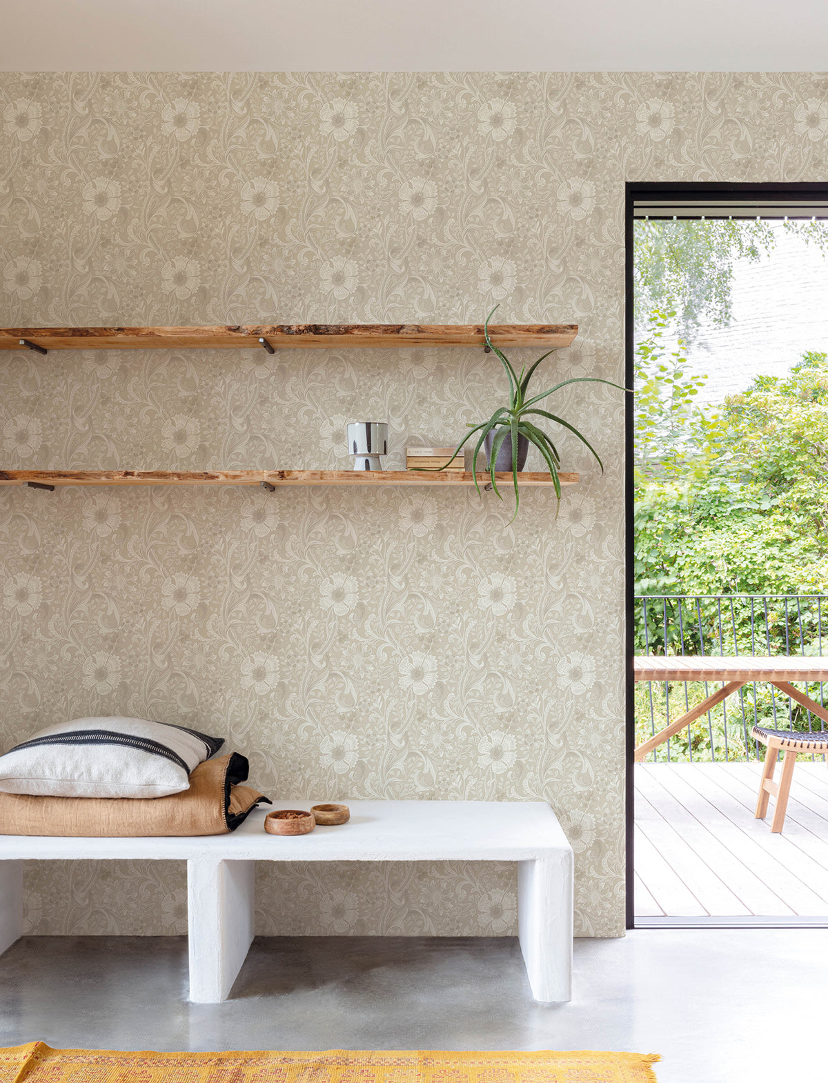 A minimalist room featuring a white bench with cushions and a blanket, wooden shelves displaying a plant and cup, and Chesapeake Liora Floral Trail Wallpaper by York Wallcoverings, with an open door revealing a green outdoor area furnished with a wooden table and chairs.