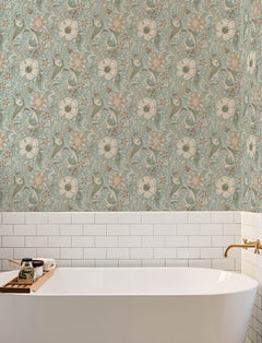 A white bathtub with a wooden tray sits against a wall adorned with York Wallcoverings’ Chesapeake Liora Floral Trail Wallpaper in green and beige, complemented by white subway tiles. A gold faucet is positioned on the right side of the tub.