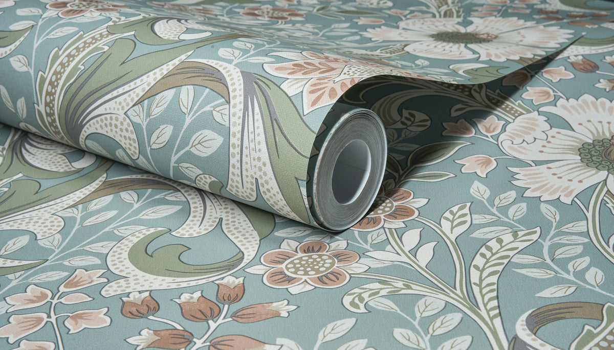 A close-up of the Chesapeake Liora Floral Trail Wallpaper by York Wallcoverings, showing the roll partially unrolled on a matching surface and highlighting its blue, green, white, and beige floral and leaf pattern.