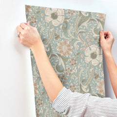 A person applies Chesapeake Liora Floral Trail Wallpaper from York Wallcoverings, featuring beige and green floral patterns, to a white wall; only their hands and striped shirt sleeves are visible.