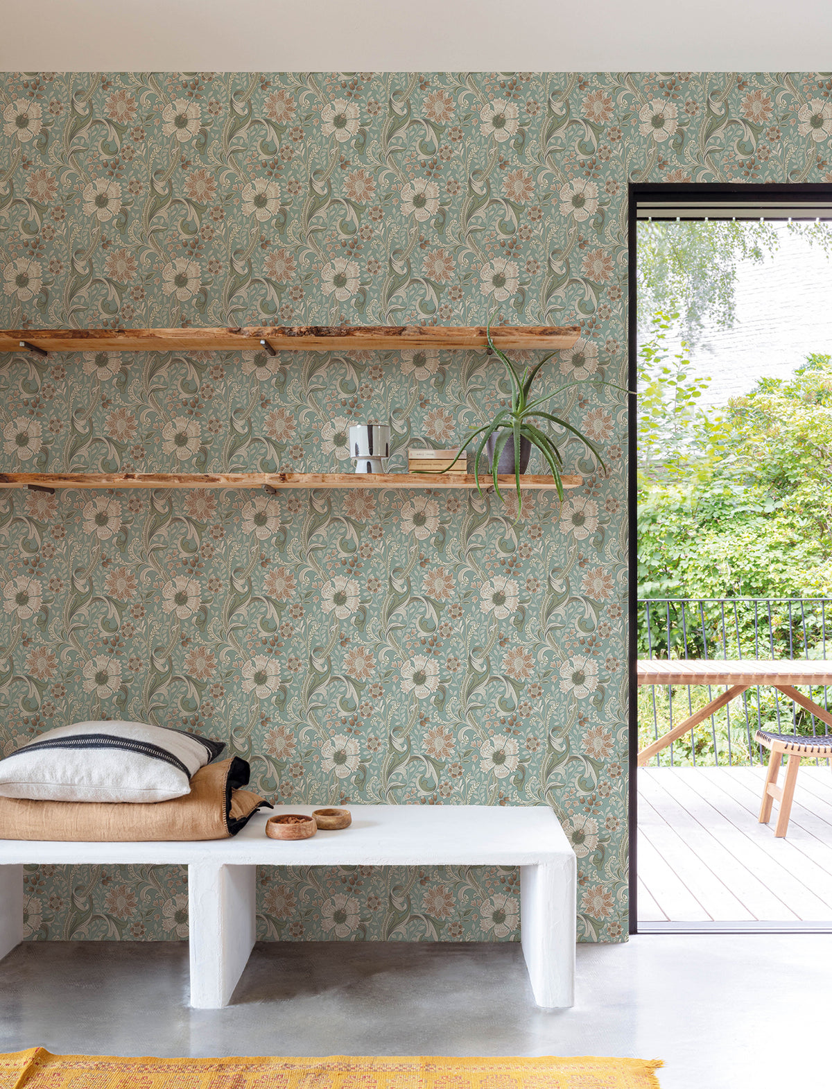 A room featuring Chesapeake Liora Floral Trail Wallpaper by York Wallcoverings, two wooden wall shelves holding books and a plant, a bench with pillows, a tray, and an open door leading to a green outdoor patio with a wooden table and bench.