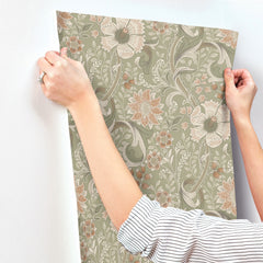 A person with a ring on their finger applies Chesapeake Liora Floral Trail Wallpaper by York Wallcoverings, featuring green and beige floral patterns, to a white wall. Only their arms and hands are visible.