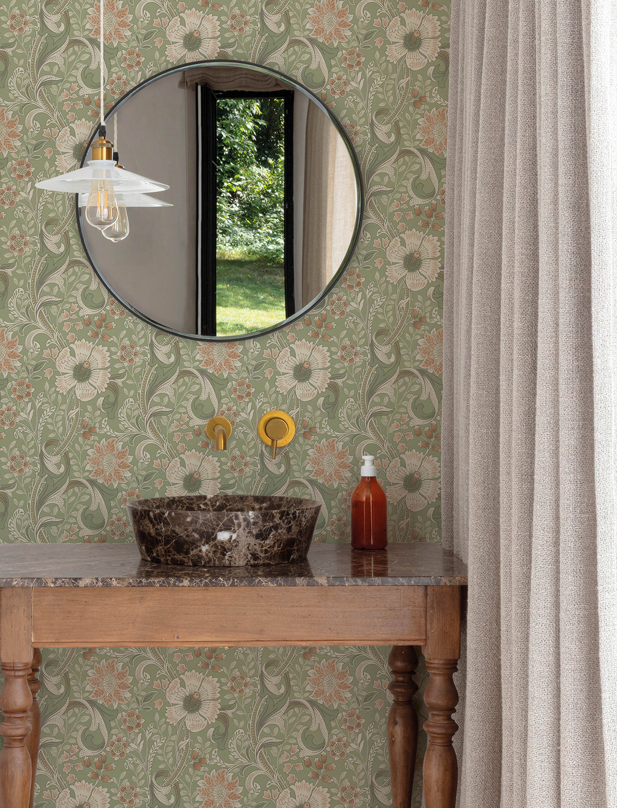 A round mirror above a marble sink, York Wallcoverings’ Chesapeake Liora Floral Trail Wallpaper, brass faucet, soap dispenser, and a hanging light fixture; beige curtain at the side and greenery visible through a window.