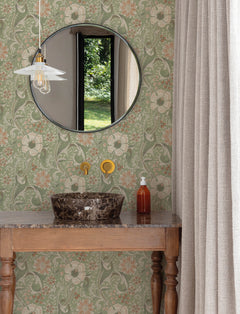 A round mirror hangs above a marble sink with gold fixtures on a wooden vanity. The York Wallcoverings Chesapeake Liora Floral Trail Wallpaper decorates the walls, complemented by beige curtains. A soap dispenser and pendant light are also present, while a window reveals greenery outside.