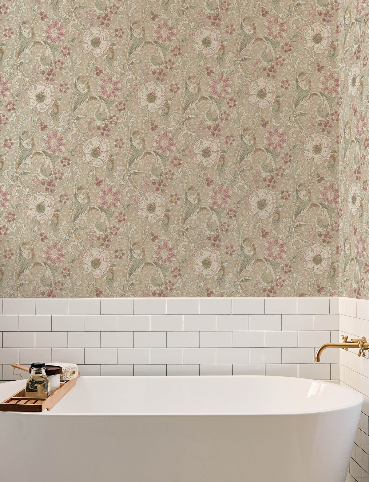 A white bathtub with a gold faucet sits against a wall adorned with York Wallcoverings’ Chesapeake Liora Floral Trail Wallpaper in beige, green, and pink, complemented by classic white subway tiles. A wooden tray on the tub holds soap and bottles.