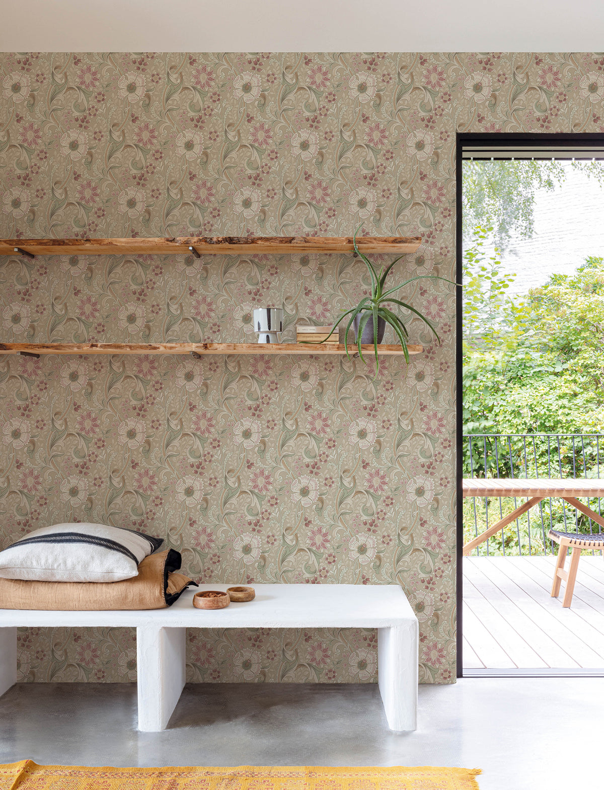 A minimalist room featuring Chesapeake Liora Floral Trail Wallpaper by York Wallcoverings, wooden shelves, a plant, books, a white bench with cushions, and an open door leading to a green outdoor area with a wooden table.