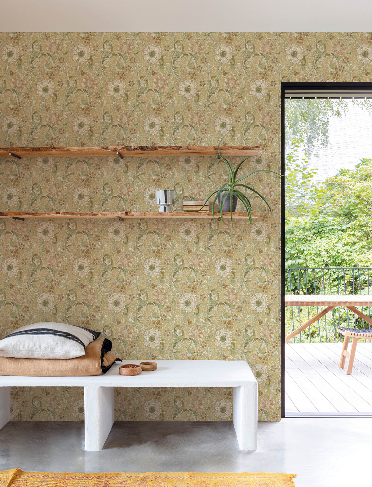 A minimalist room featuring York Wallcoverings’ Chesapeake Liora Floral Trail Wallpaper, three wooden shelves, a white bench with cushions, and a potted plant. A glass door opens to a green outdoor patio with a wooden table and bench.